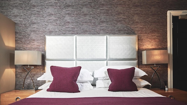 A neatly made bed with a white headboard, two red pillows, and a red throw blanket. There are two bedside tables with lamps and a wall with a textured pattern. - Great Fosters (Egham)