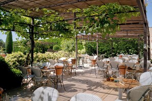 Un espace extérieur aménagé pour les repas, avec des tables et des chaises, et recouvert d’une pergola ornée de plantes grimpantes.