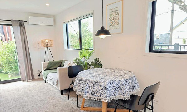 Um quarto aconchegante com uma cama, uma mesa redonda com toalha de mesa estampada, um abajur e uma grande janela com vista para o verde.