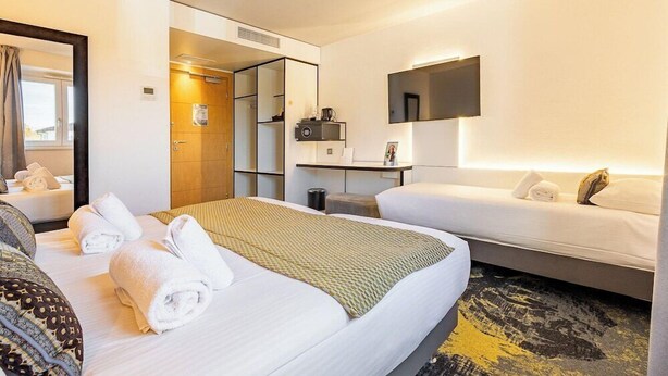 A hotel room with two beds, a flat-screen TV, a mirror, and a small table with a lamp. - Brit Hotel Le Relais du Ried (Bischwihr)