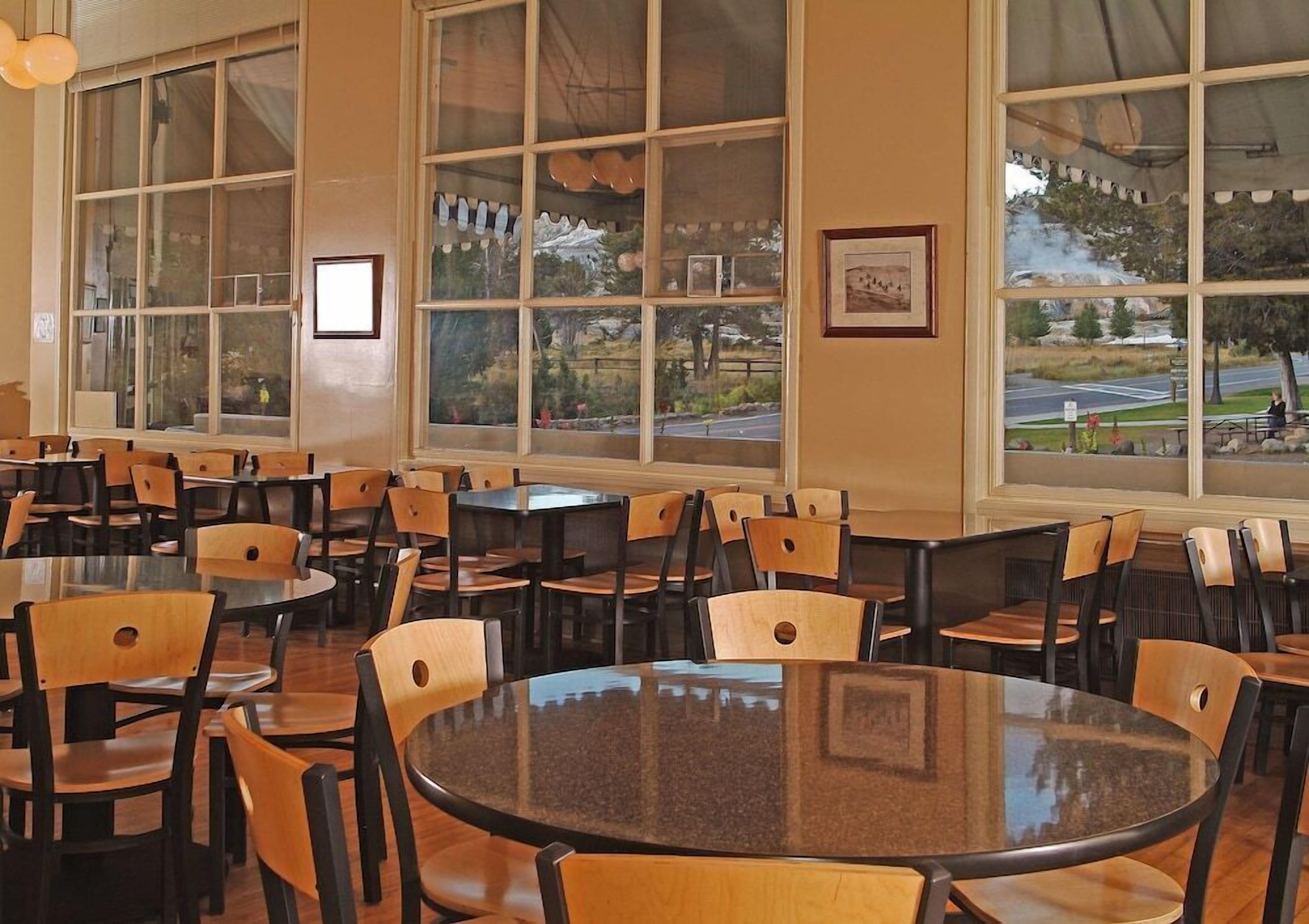 A spacious dining area with multiple tables and chairs, large windows with a view of a park and road, and framed pictures on the walls.