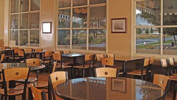 A spacious dining area with multiple tables and chairs, large windows with a view of a park and road, and framed pictures on the walls.