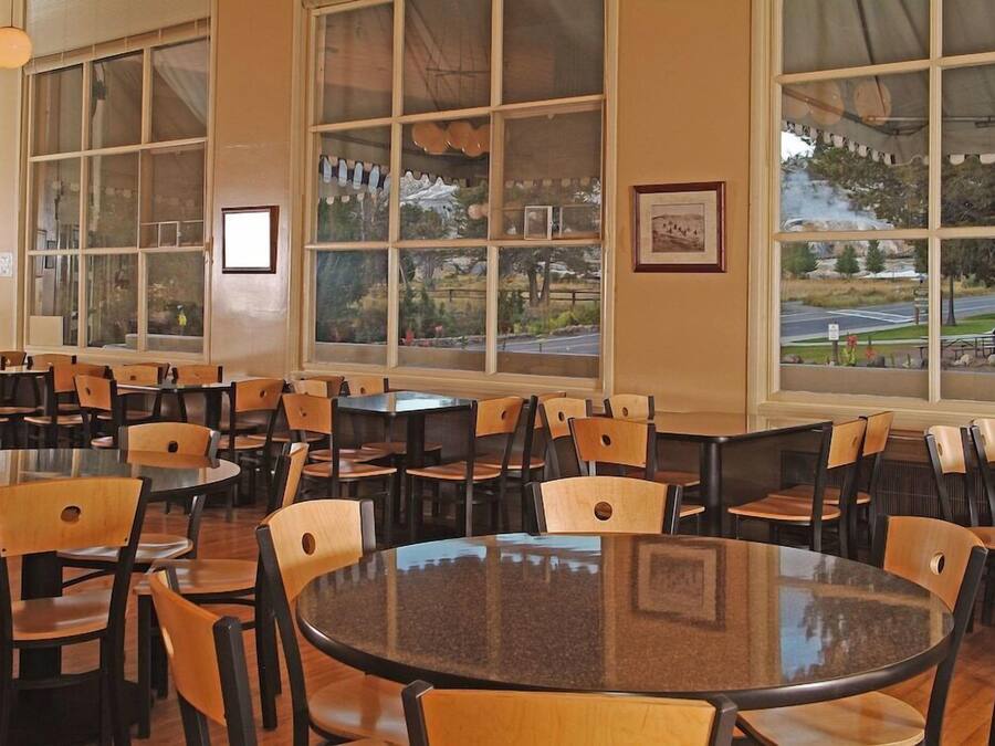 A spacious dining area with multiple tables and chairs, large windows with a view of a park and road, and framed pictures on the walls.