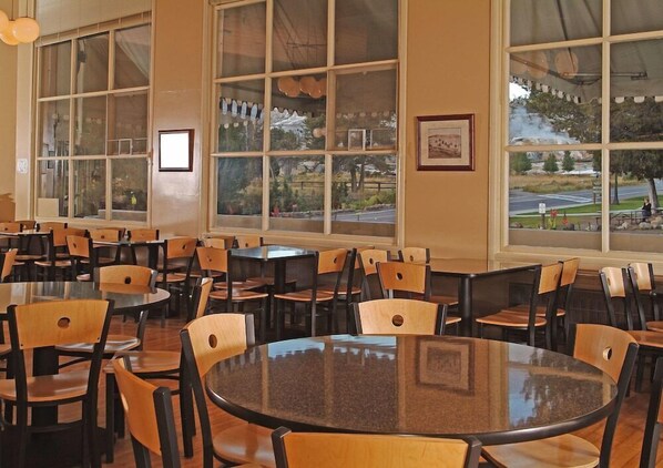 Un comedor amplio con varias mesas y sillas, ventanas grandes con vistas a un parque y una calle, y cuadros en las paredes.