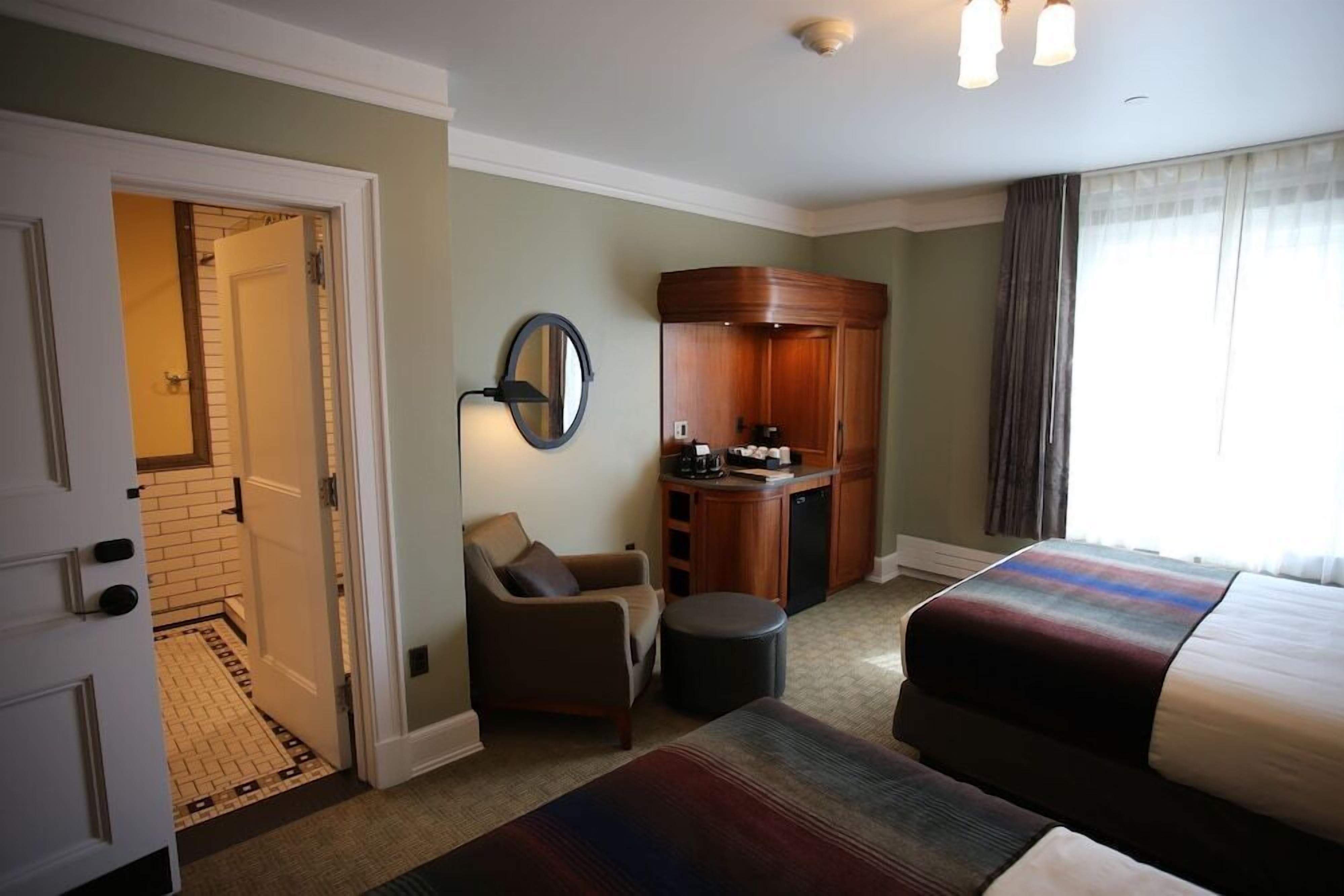 Une chambre d’hôtel comprenant un lit, une chaise, une petite table, un miroir, un bureau avec une machine à café et une vue sur l’extérieur par une fenêtre.
