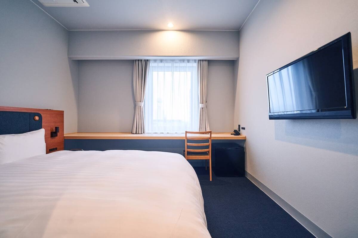 Ett hotellrum med en säng, ett skrivbord, en stol, en tv och ett fönster med gardiner.