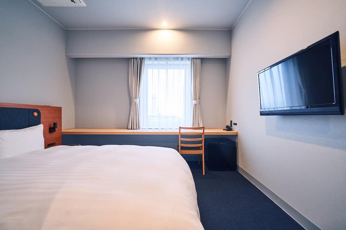 A hotel room with a bed, a desk, a chair, a TV, and a window with curtains.