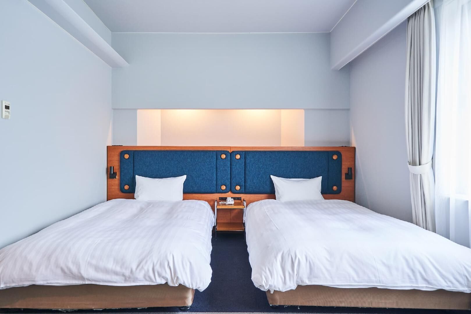 Ein Hotelzimmer mit zwei Betten, einem Holzkopfteil, einem Nachttisch und einem Fenster mit Vorhängen.