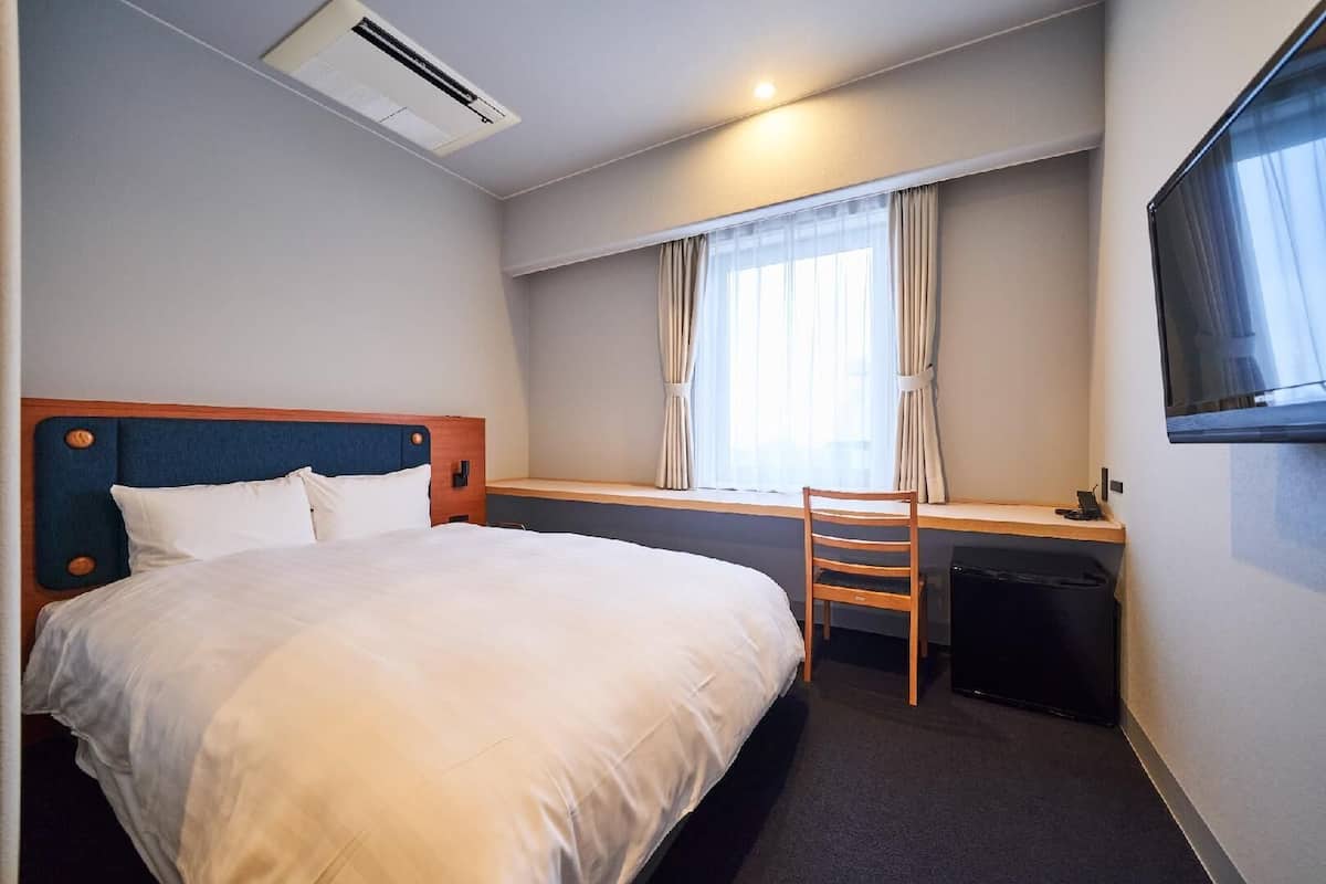 Ett hotellrum med en säng, ett skrivbord, en stol, en luftkonditioneringsanläggning, ett fönster med gardiner och en tv som är monterad på väggen.