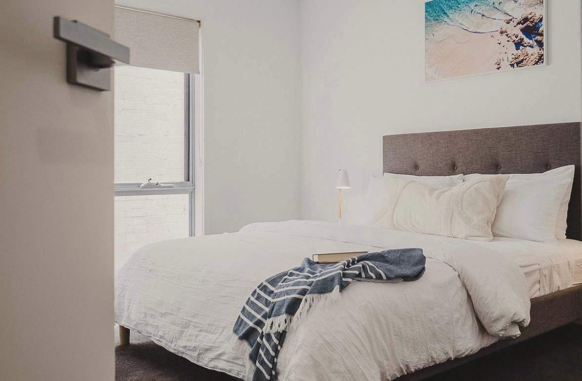 A neatly made bed with a gray headboard, a window with a view, and a painting on the wall.