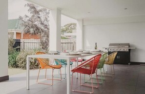 Un área de comedor al aire libre con una mesa larga y sillas coloridas, una parrilla y vistas a un jardín y una casa al fondo.