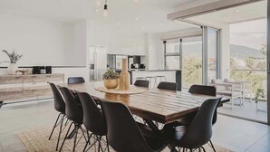 A modern dining area with a wooden table and black chairs, a kitchen in the background, and a view of the outdoors.