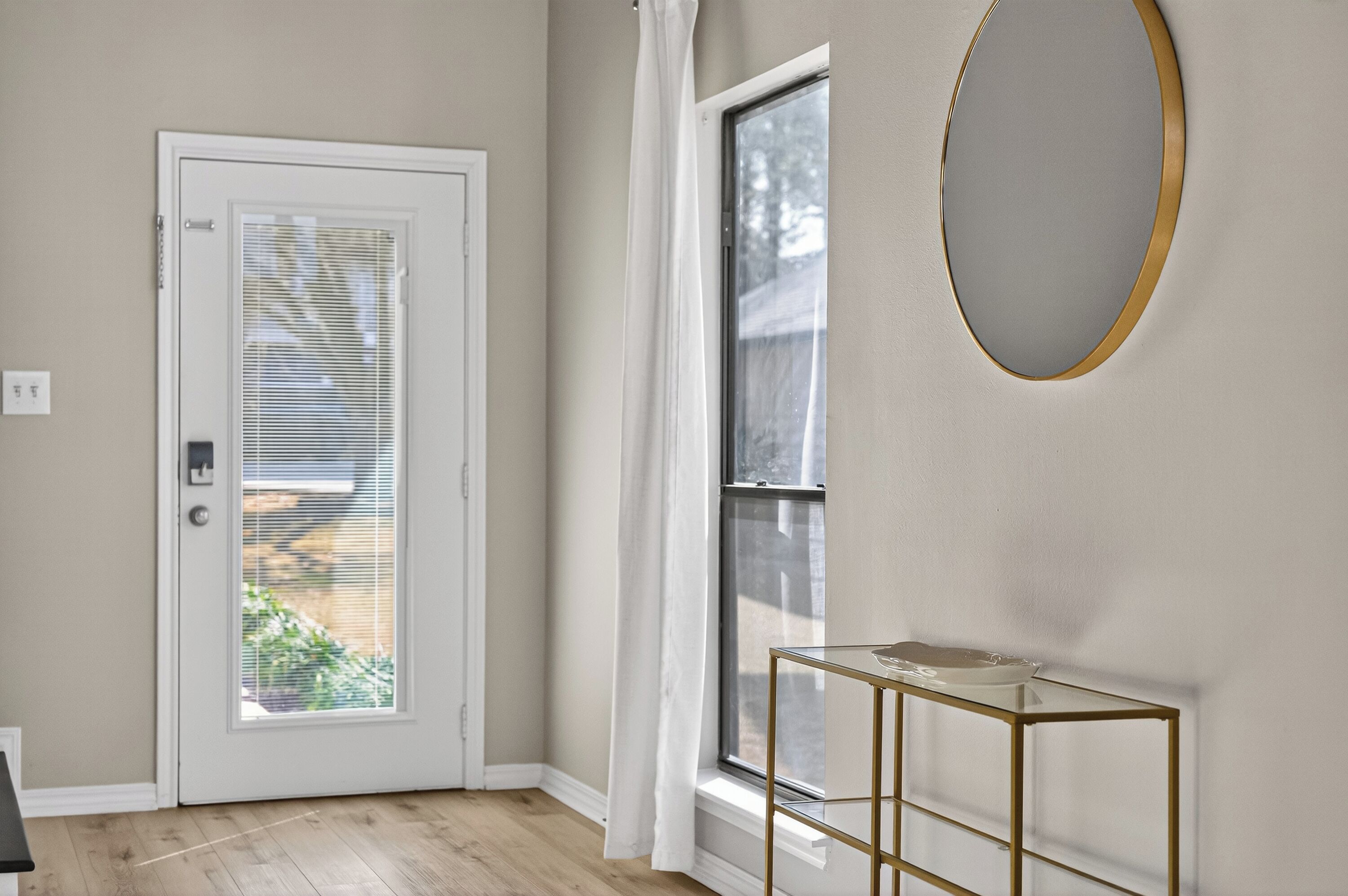 A room with a white door, a window with blinds, a round mirror, and a glass table.