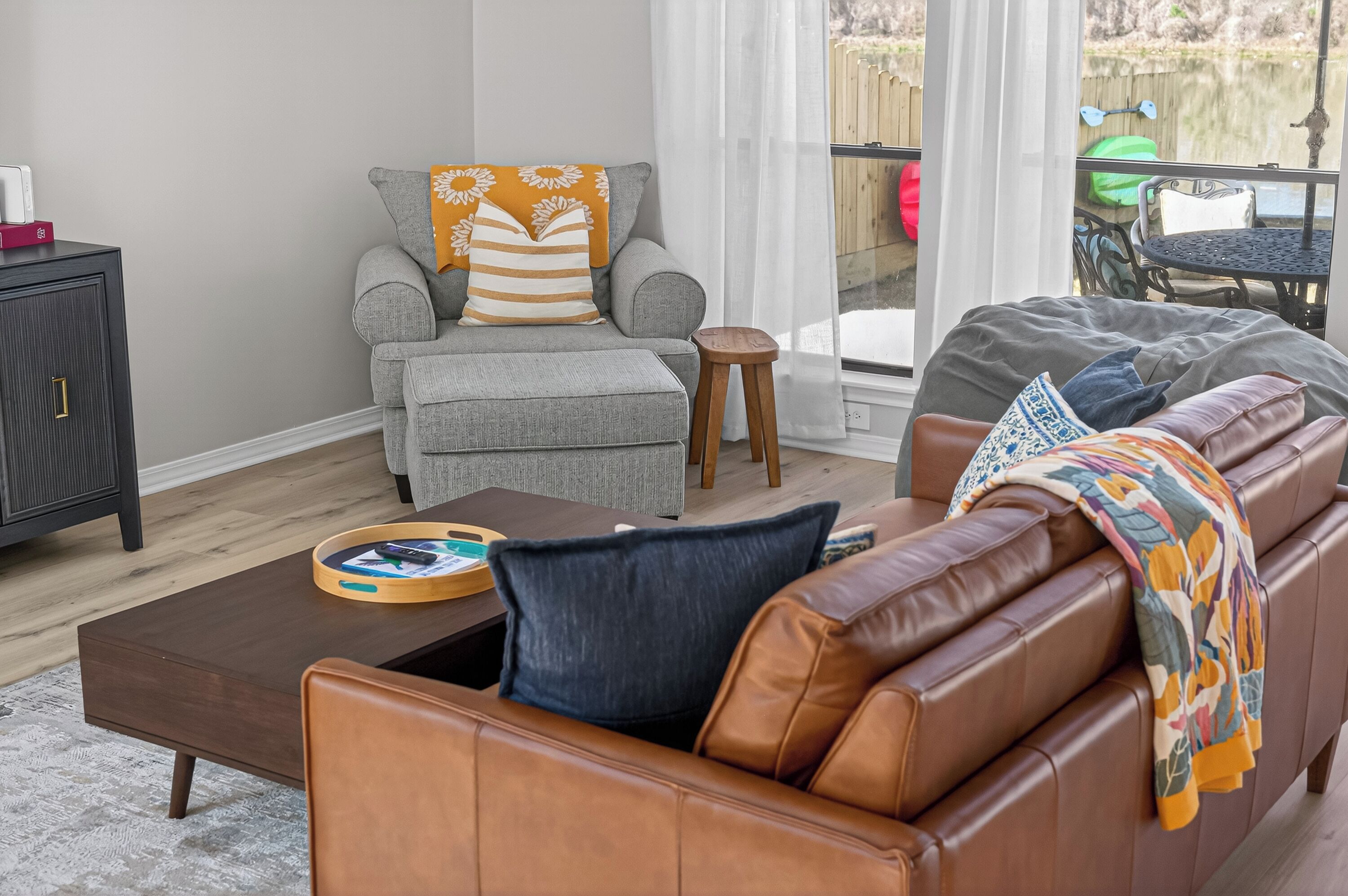 Un salon avec un canapé en cuir marron, un fauteuil gris agrémenté d’un coussin à rayures, une table basse en bois et une porte-fenêtre coulissante donnant sur un espace extérieur.