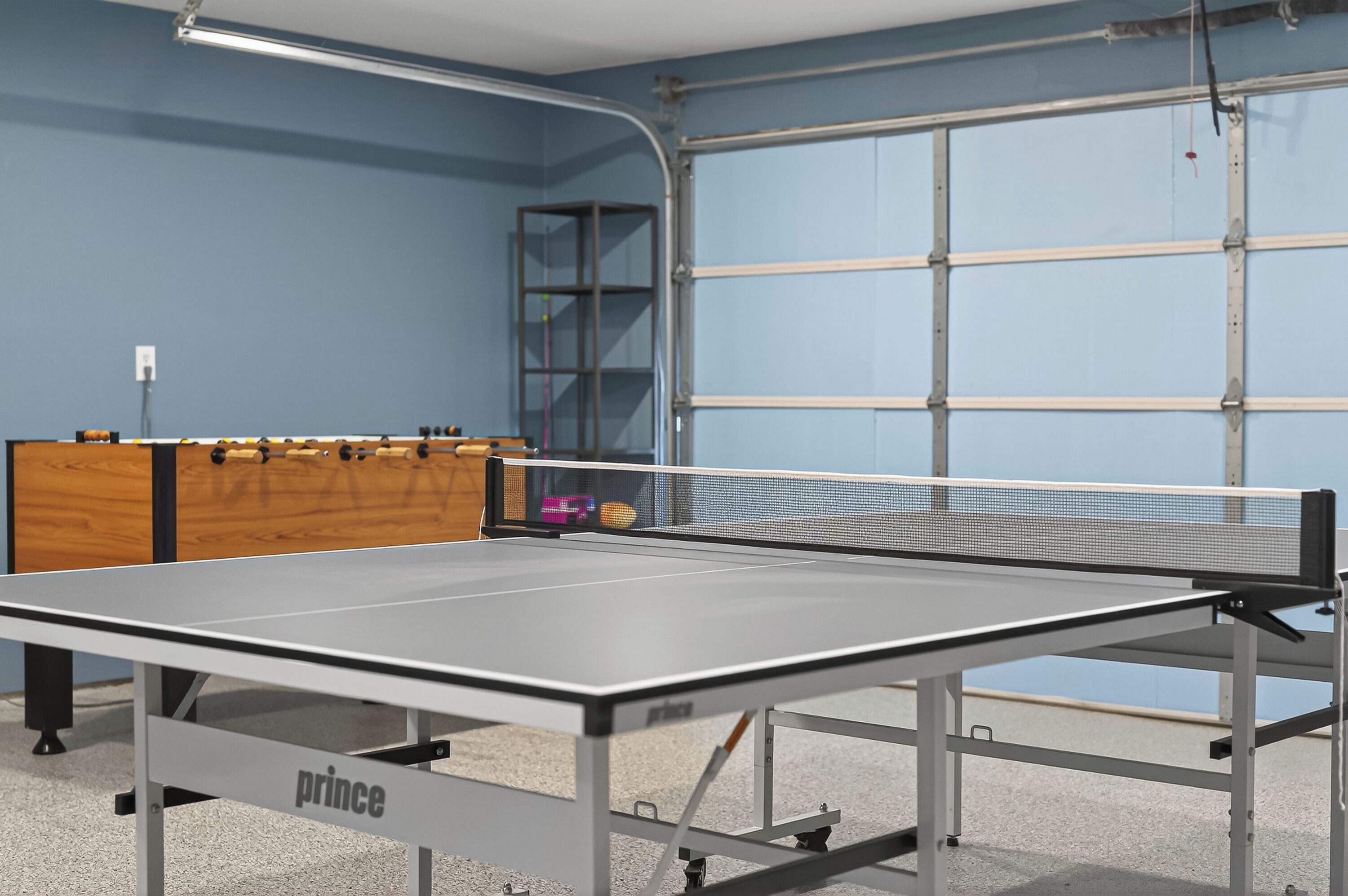 Ein Tischtennis-Tisch mit Netz, eine Holzschrank mit Gegenständen und eine blaue Wand mit einer Metalldür.'