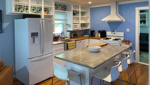 A compact kitchen with white appliances, a central island with bar stools, and a view of the adjacent living area. - Crabapple Cottage (Essex)