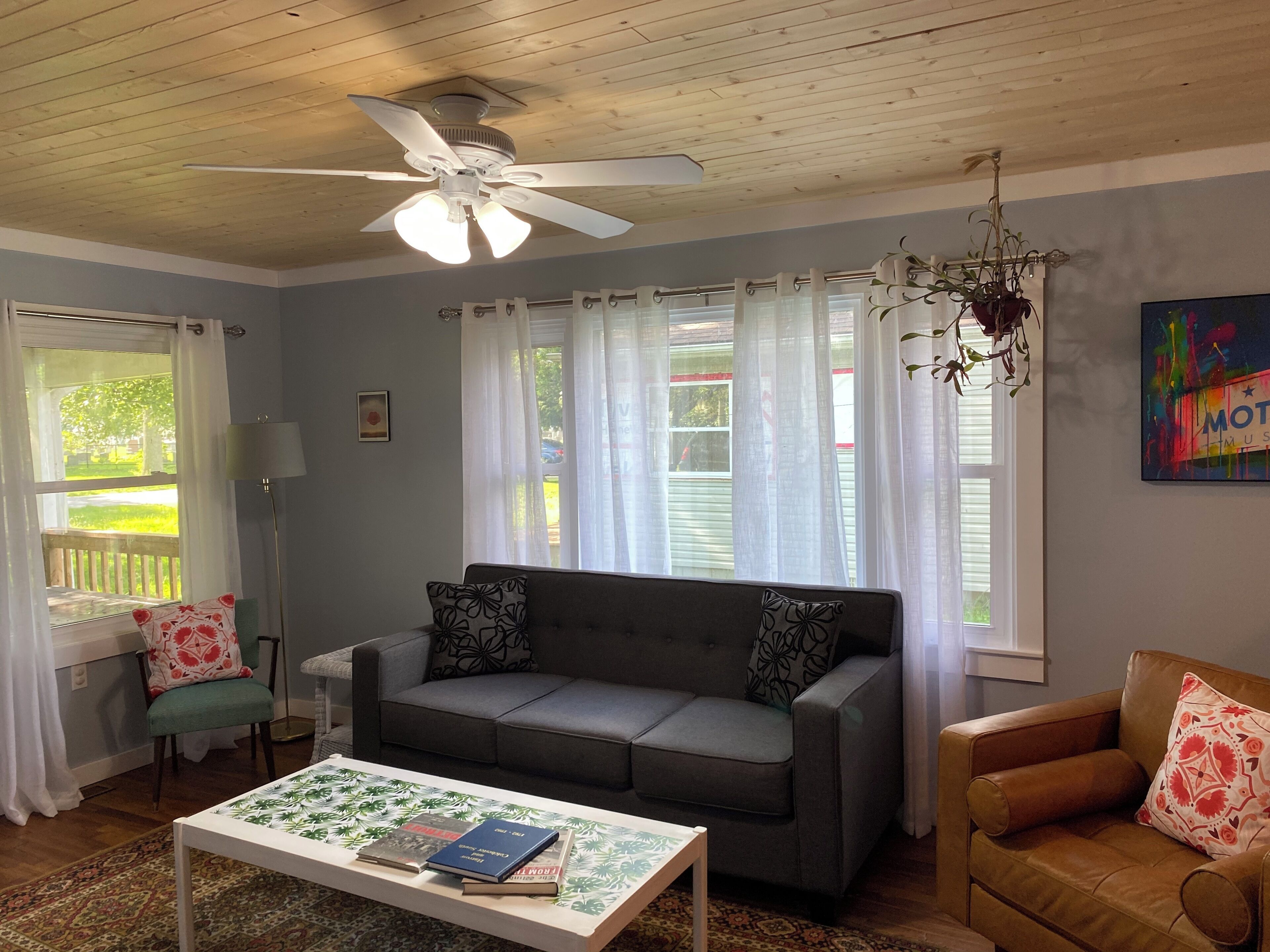 A living room with a grey sofa, a brown leather armchair, a white coffee table with books, a ceiling fan, and a window with sheer curtains.