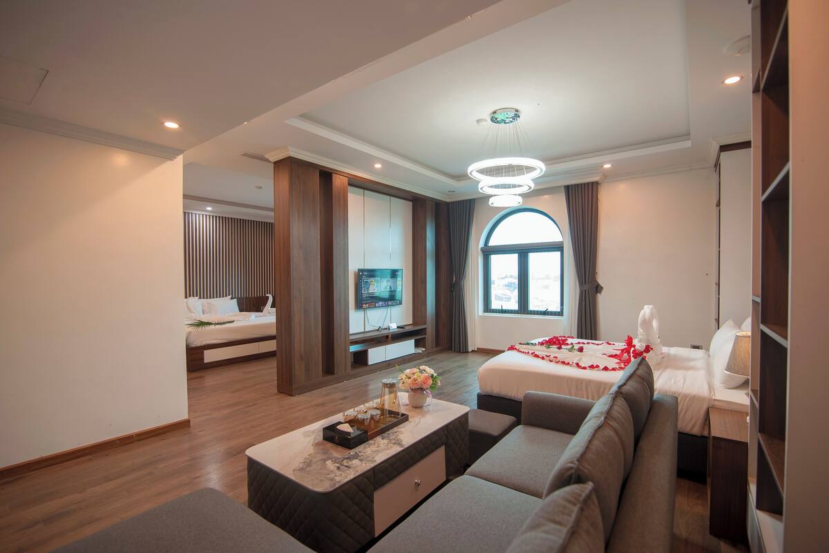 A modern hotel room with a sofa, a TV, a bed with a red rose decoration, and a large window.