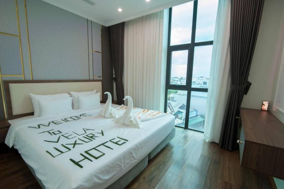 A hotel room with a large bed, a view of the city through a window, and a desk with a lamp.