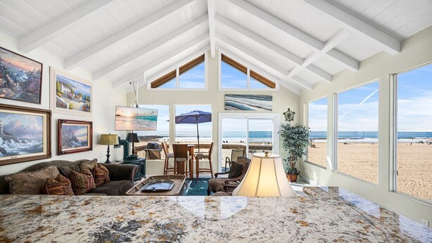 A spacious living room with a large marble floor, a high ceiling with exposed beams, and large windows offering a beach view.