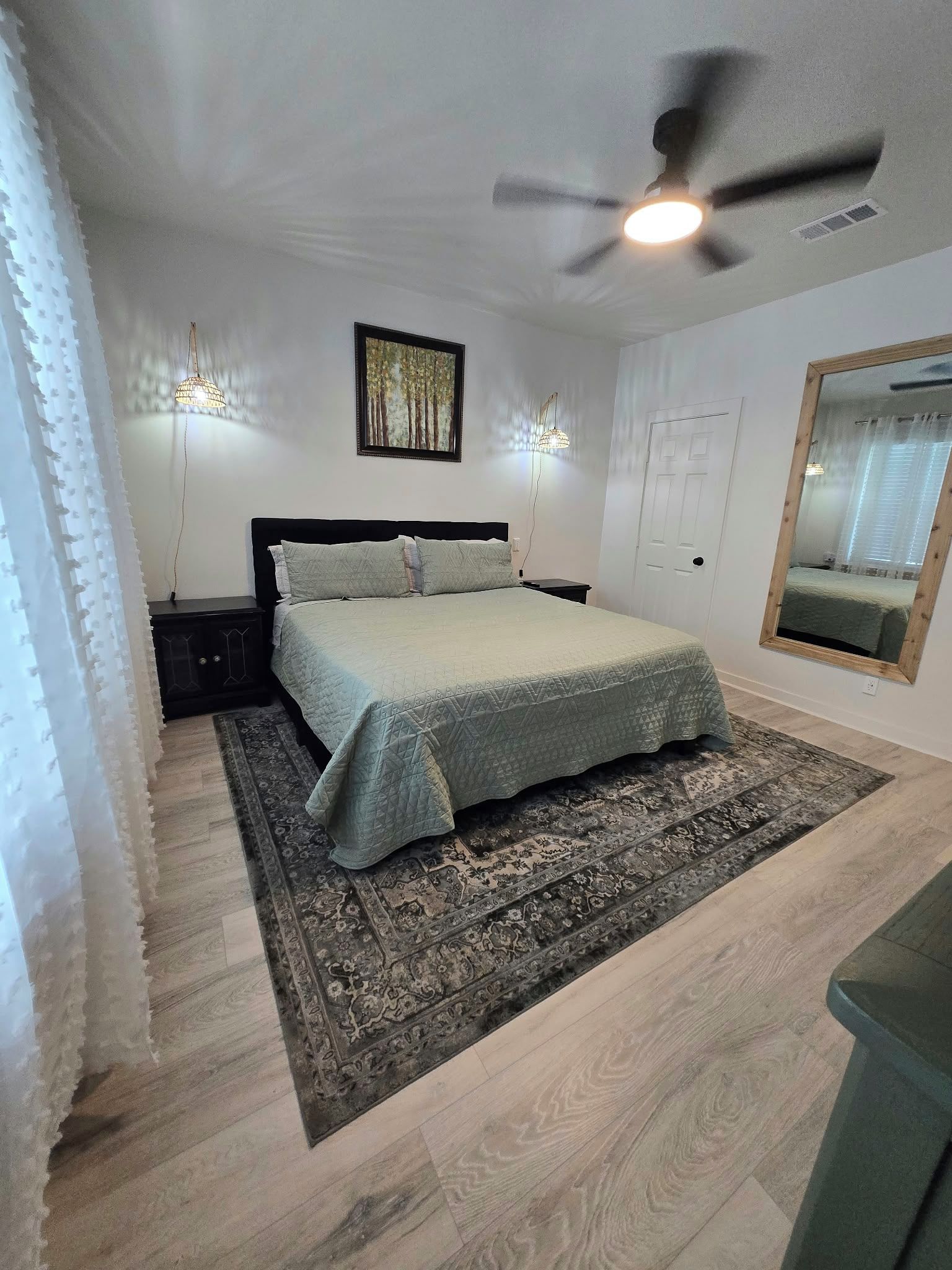 A bedroom with a bed, a ceiling fan, a wall-mounted light, a door, and a mirror.