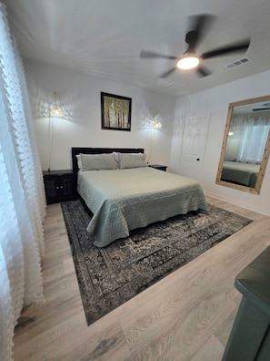 Un dormitorio con una cama, ventilador de techo, lámpara de pared, una puerta y un espejo.