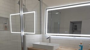A modern bathroom with a glass shower enclosure, a white sink, wooden cabinets, and a large mirror with integrated lighting. - Twin Nest Duplex (Irving)