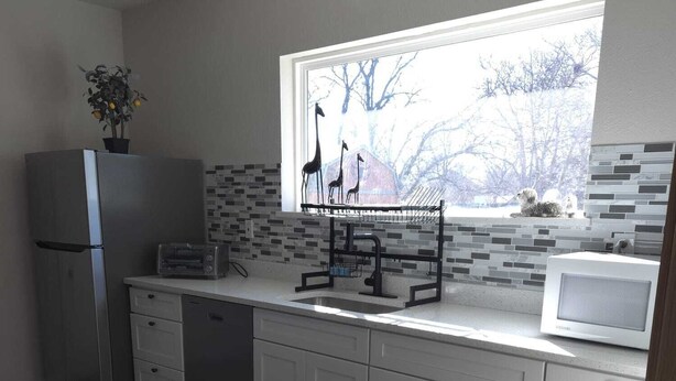 A modern kitchen with white cabinets, a stainless steel refrigerator, a microwave, and a window with a view of trees. - Twin Nest Duplex (Irving)