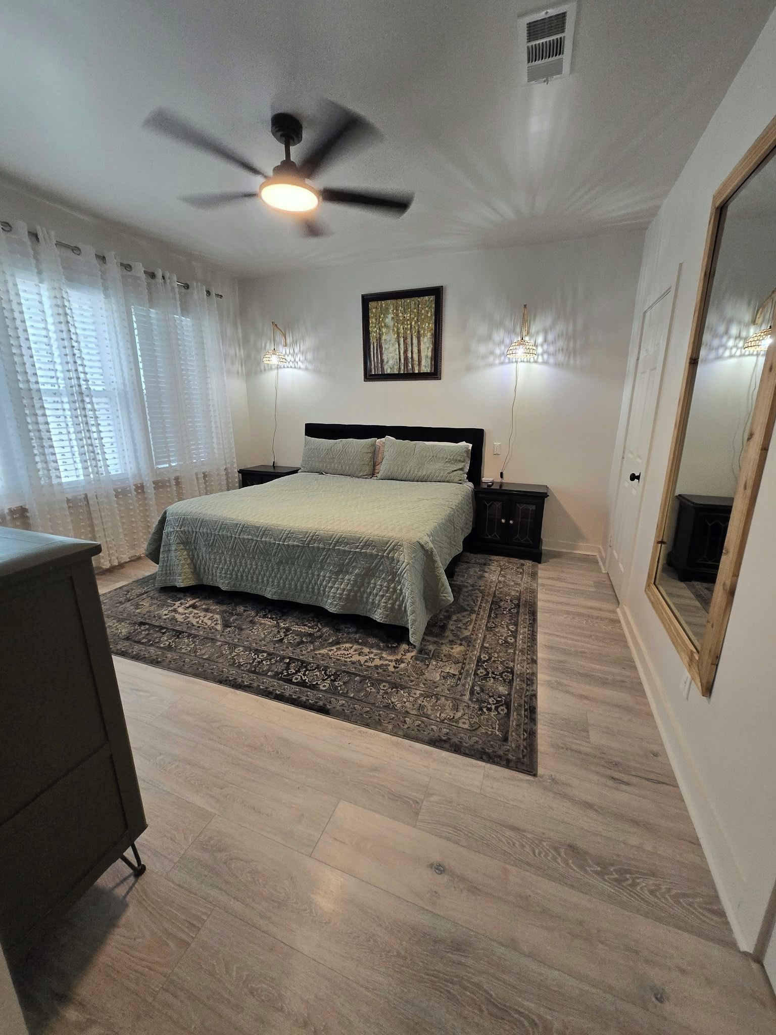 Un dormitorio con una cama, ventilador de techo, mesita de noche, cómoda, alfombra y espejo.