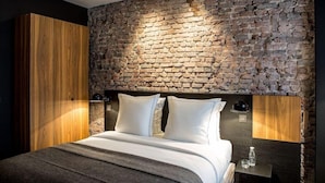 A modern bedroom with a stone wall, a wooden wardrobe, a bed with white bedding, and a bedside table with a bottle and glass. - Hotel Louvre Lens (Lens)