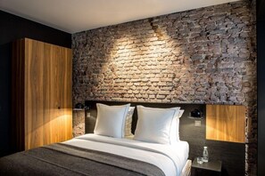 A modern bedroom with a stone wall, a wooden wardrobe, a bed with white bedding, and a bedside table with a bottle and glass. - Hotel Louvre Lens (Lens)