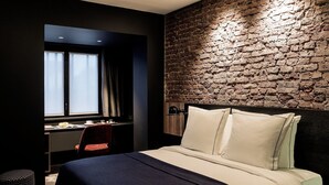 A modern hotel room with a brick wall, a large bed with white linens, a desk with a chair, and a window with curtains. - Hotel Louvre Lens (Lens)