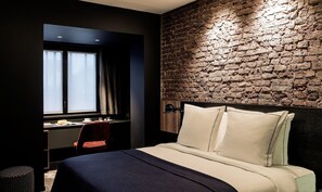A modern hotel room with a brick wall, a large bed with white linens, a desk with a chair, and a window with curtains. - Hotel Louvre Lens (Lens)