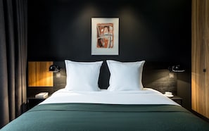 A modern hotel room with a dark wall, a framed abstract painting, a wooden headboard, two bedside tables with lamps, and a green bedspread. - Hotel Louvre Lens (Lens)
