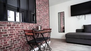 A room with a brick wall, a wooden table with a chair, a grey sofa, and a mounted TV. - Alive Hotel de Quebec (Rouen)