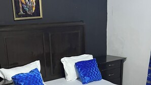 Un lit avec une literie blanche et deux oreillers bleus, une tête de lit en bois foncé, une petite table de chevet et un tapis gris sur un sol carrelé.
