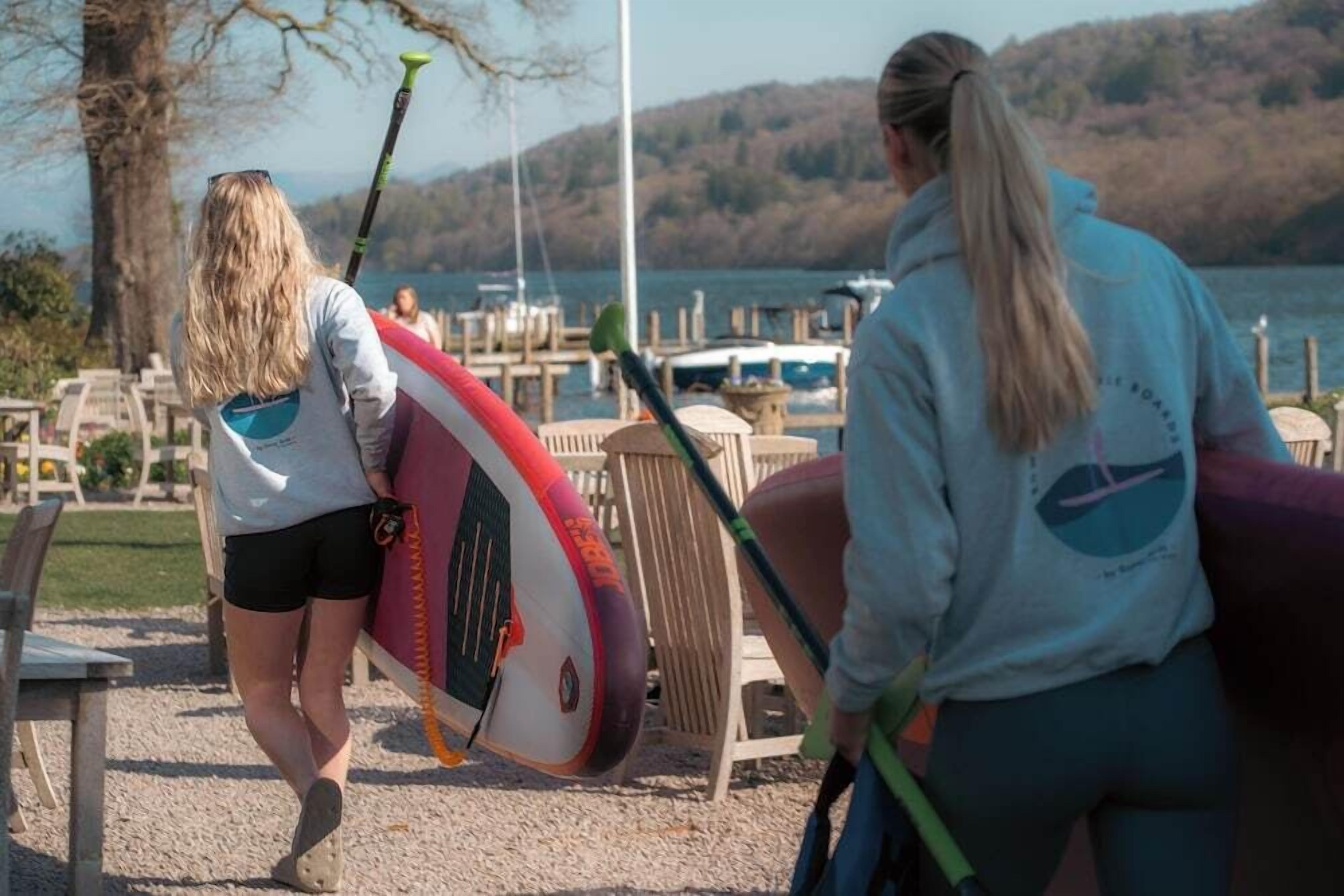 Two people carrying paddleboards near a lakeside area with boats and dock chairs.