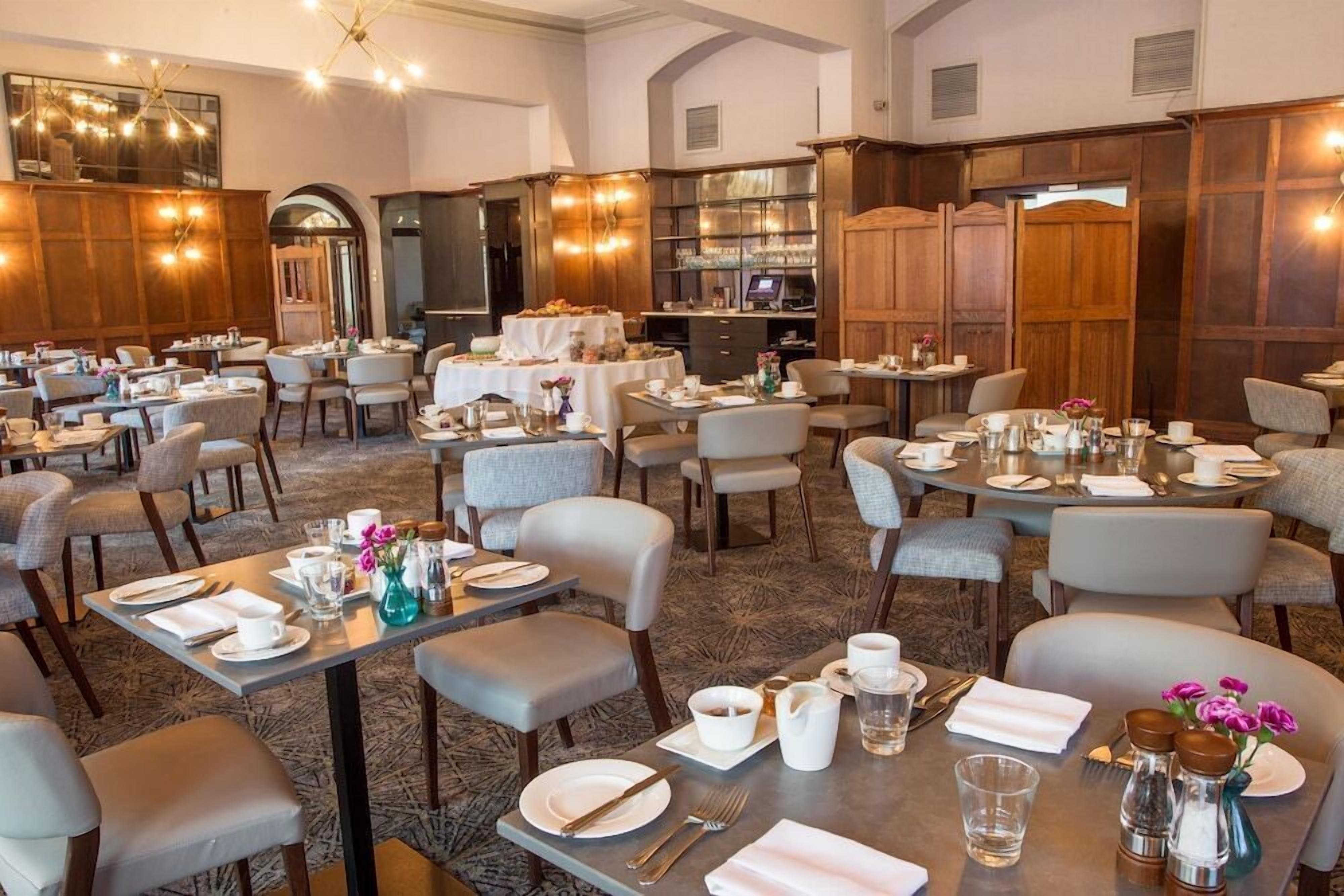Un vaste espace dédié à la restauration dans l’hôtel, avec de nombreuses tables préparées pour le petit-déjeuner, comprenant un assortiment de chaises et de tables, et offrant une atmosphère chaleureuse et accueillante grâce à un lambris en bois et un éclairage doux.