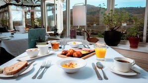 Mesa de desayuno dispuesta con croissants, huevos, jamón y jugo, con vistas panorámicas a través de amplias ventanas.