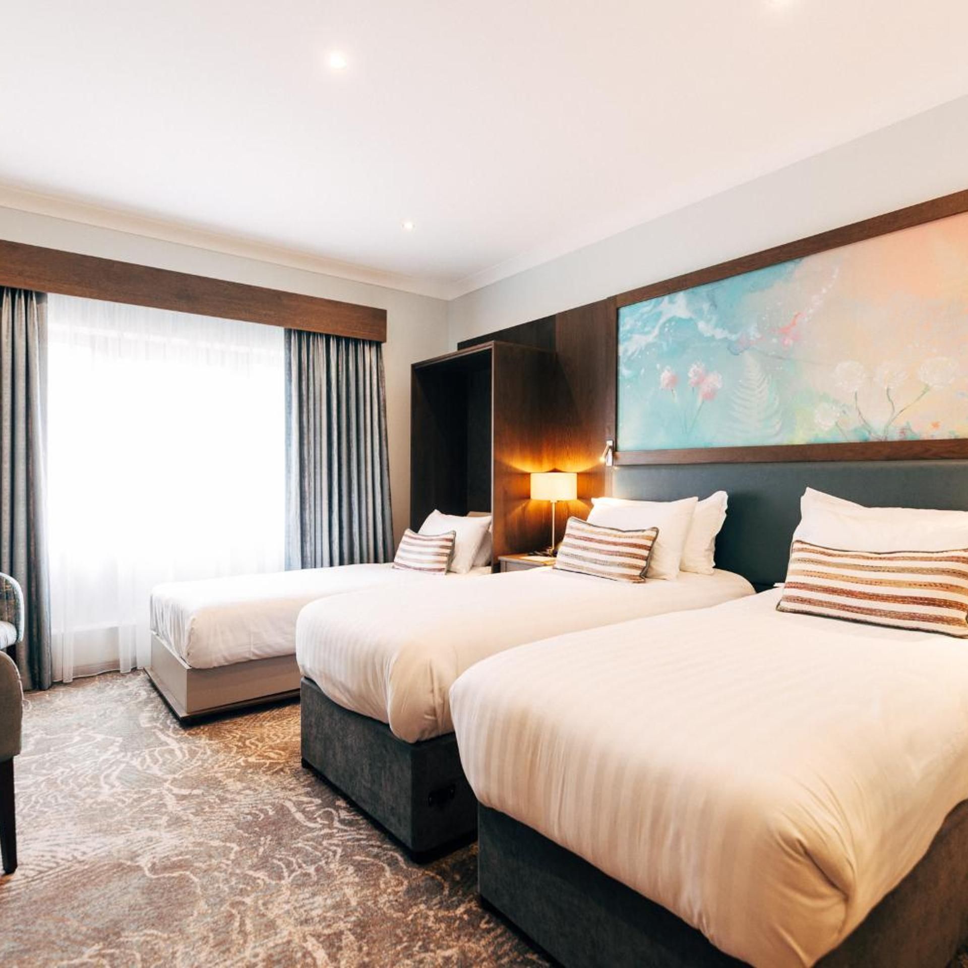 A hotel room with two beds, a large window with curtains, a painting on the wall, and a carpeted floor.