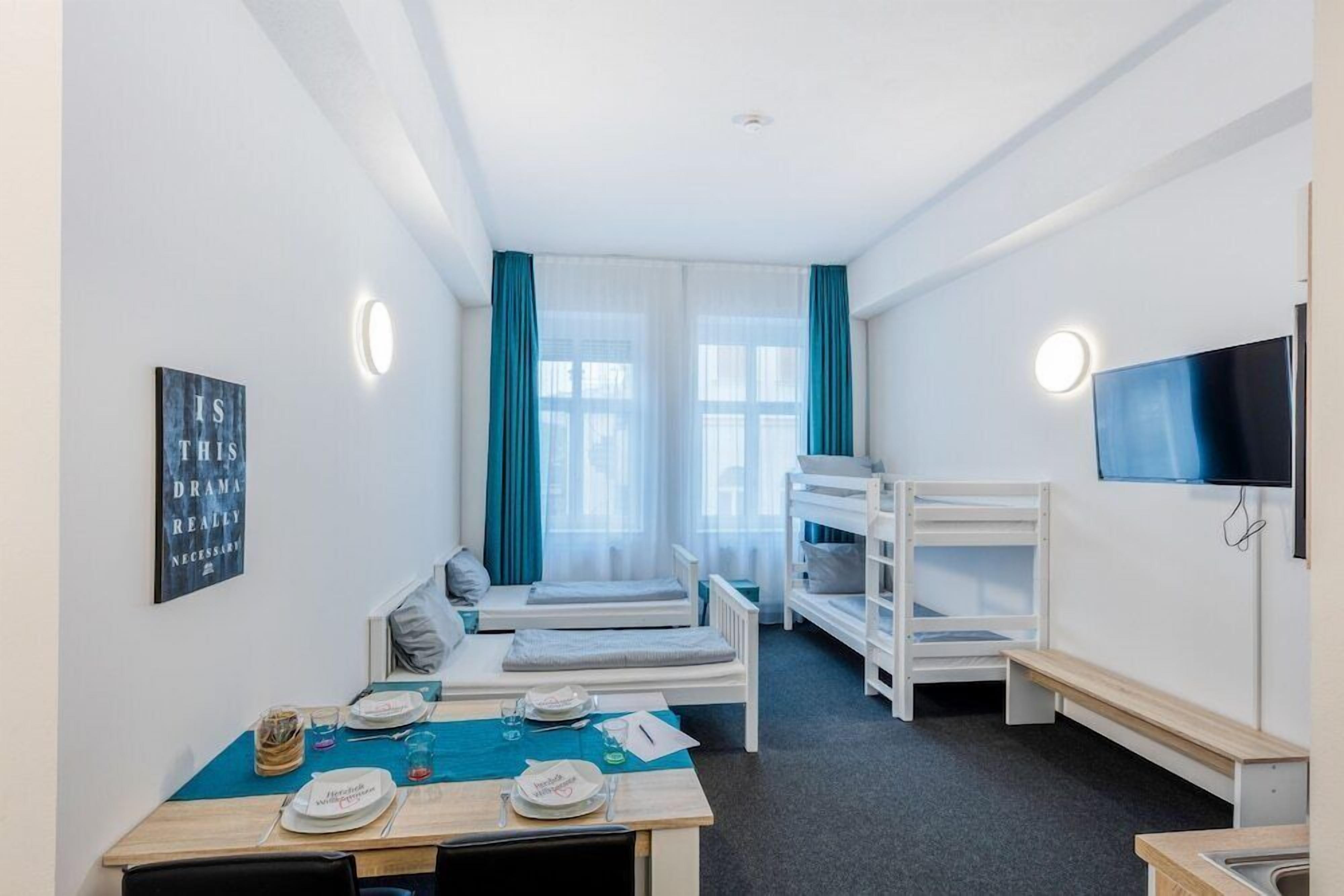 Ein Hotelzimmer mit Stockbetten, einem Esstisch gedeckt mit Tellern und Gläsern, einem an der Wand montierten Fernseher und einem Fenster mit Vorhängen.