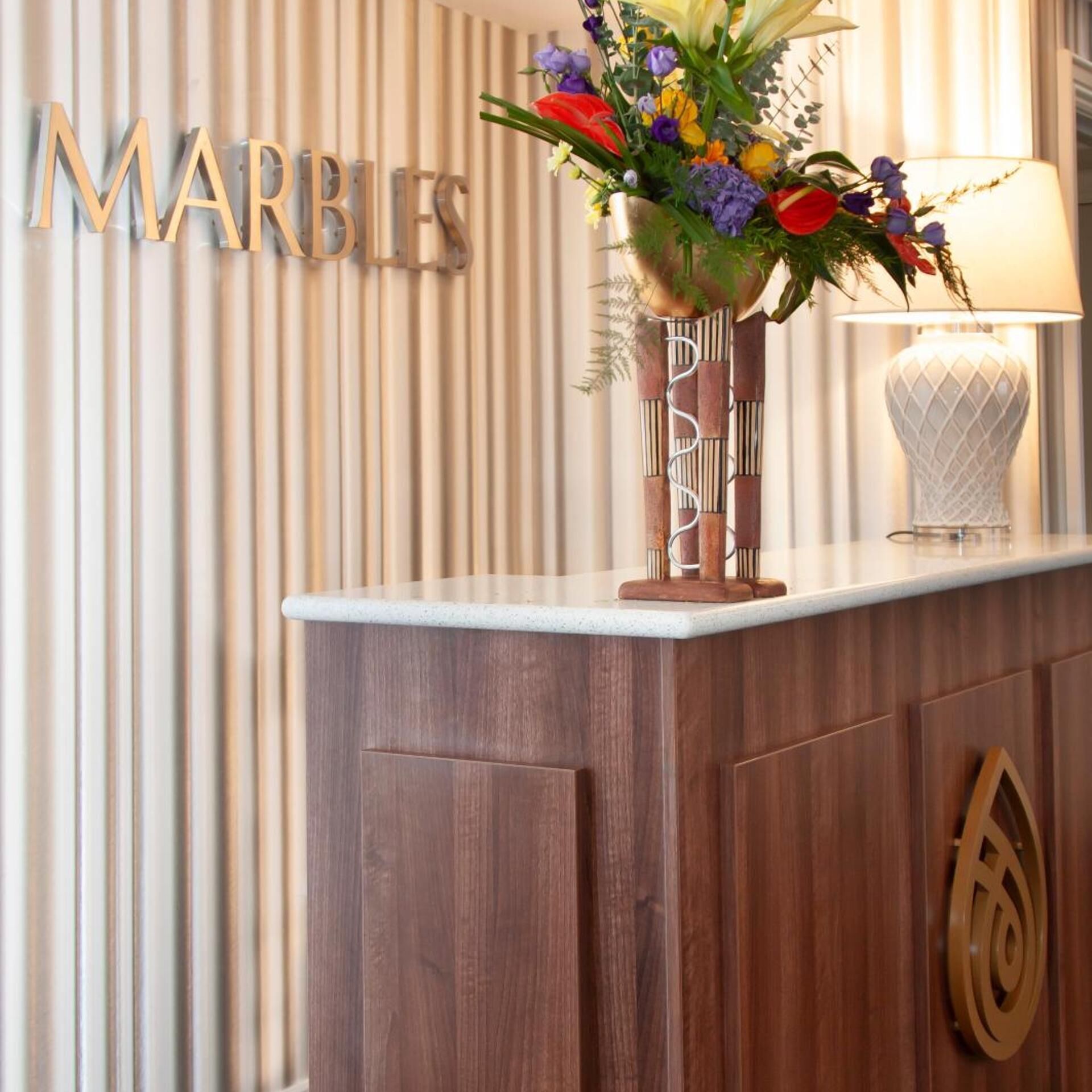 A reception desk with a vase of flowers, a lamp, and a wall with the word 'MARBLES' in large letters.