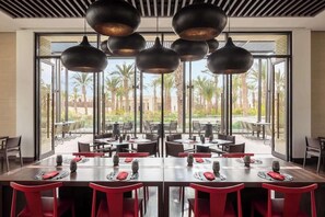 Un área de comedor moderna con varias mesas y sillas, amplias ventanas que ofrecen vista a palmeras y edificios, e iluminación colgante.