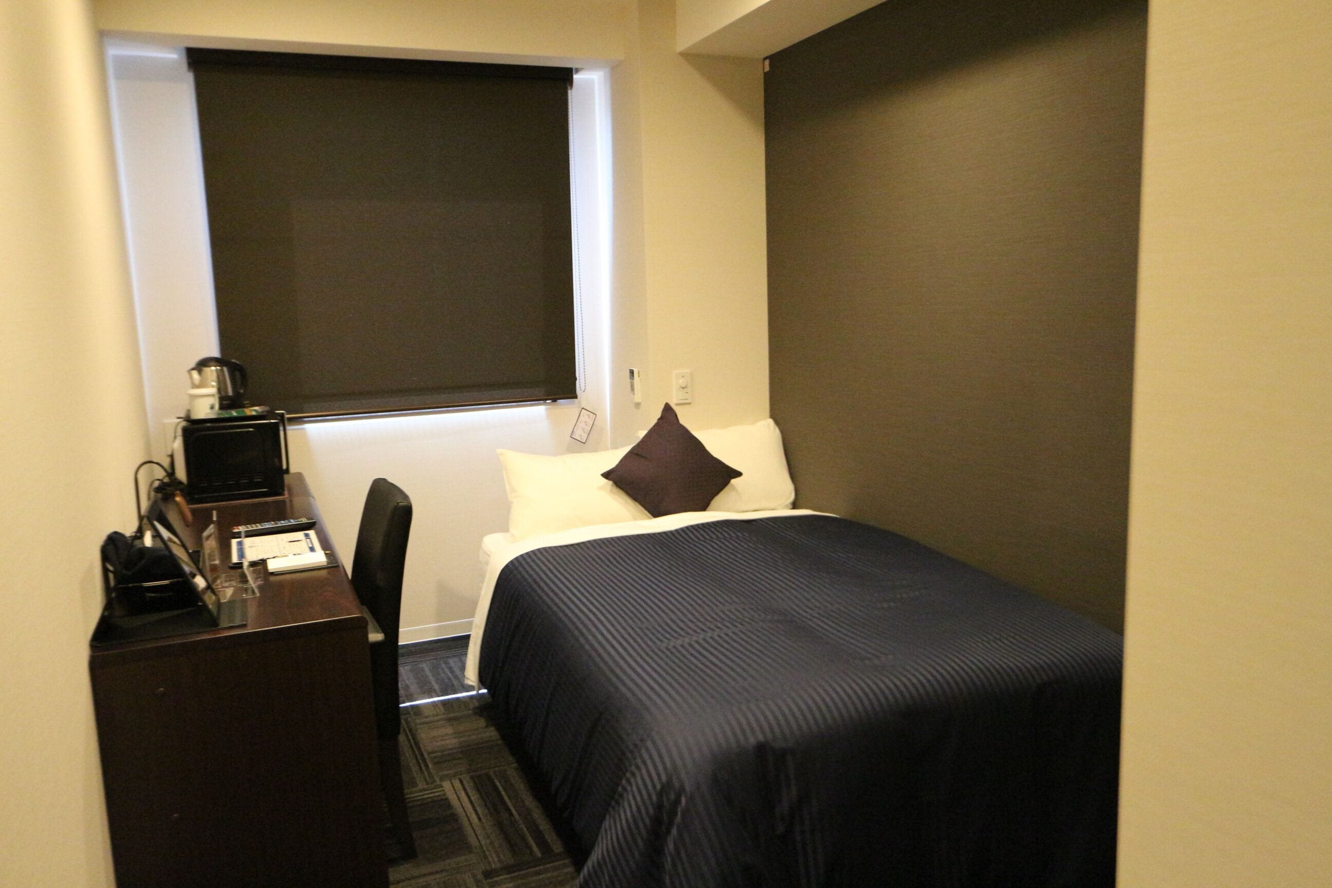 Habitación de hotel con cama, escritorio, silla, ventana con persiana y teléfono en la pared.