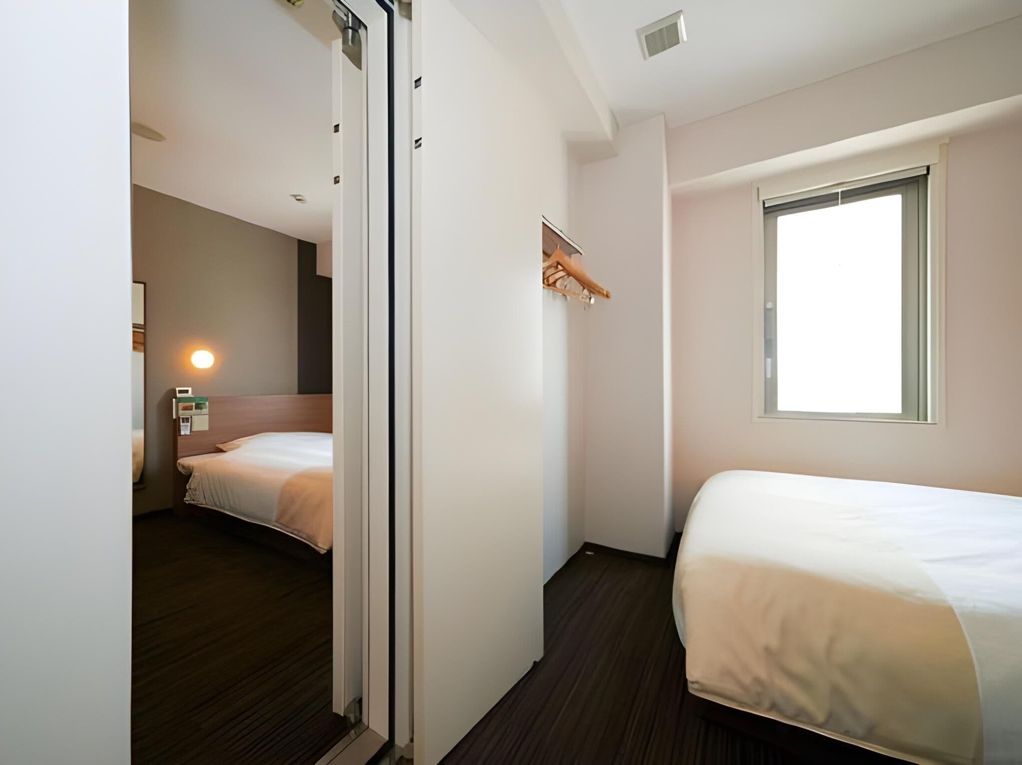 Une chambre d’hôtel avec deux lits, un plancher en bois, une fenêtre avec des stores et une étagère fixée au mur, équipée d’un porte-serviettes.