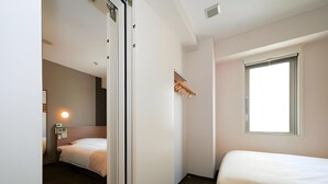 Ein Hotelzimmer mit zwei Betten, Holzboden, einem Fenster mit Jalousien und einem an der Wand befestigten Regal mit Handtuchhalter.