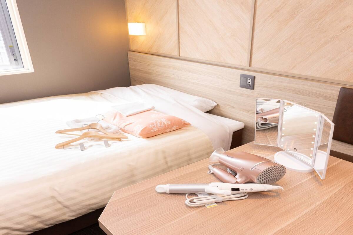 Une chambre d’hôtel comprenant un lit, une table de chevet avec un miroir de maquillage, un sèche-cheveux et un adaptateur de voyage.