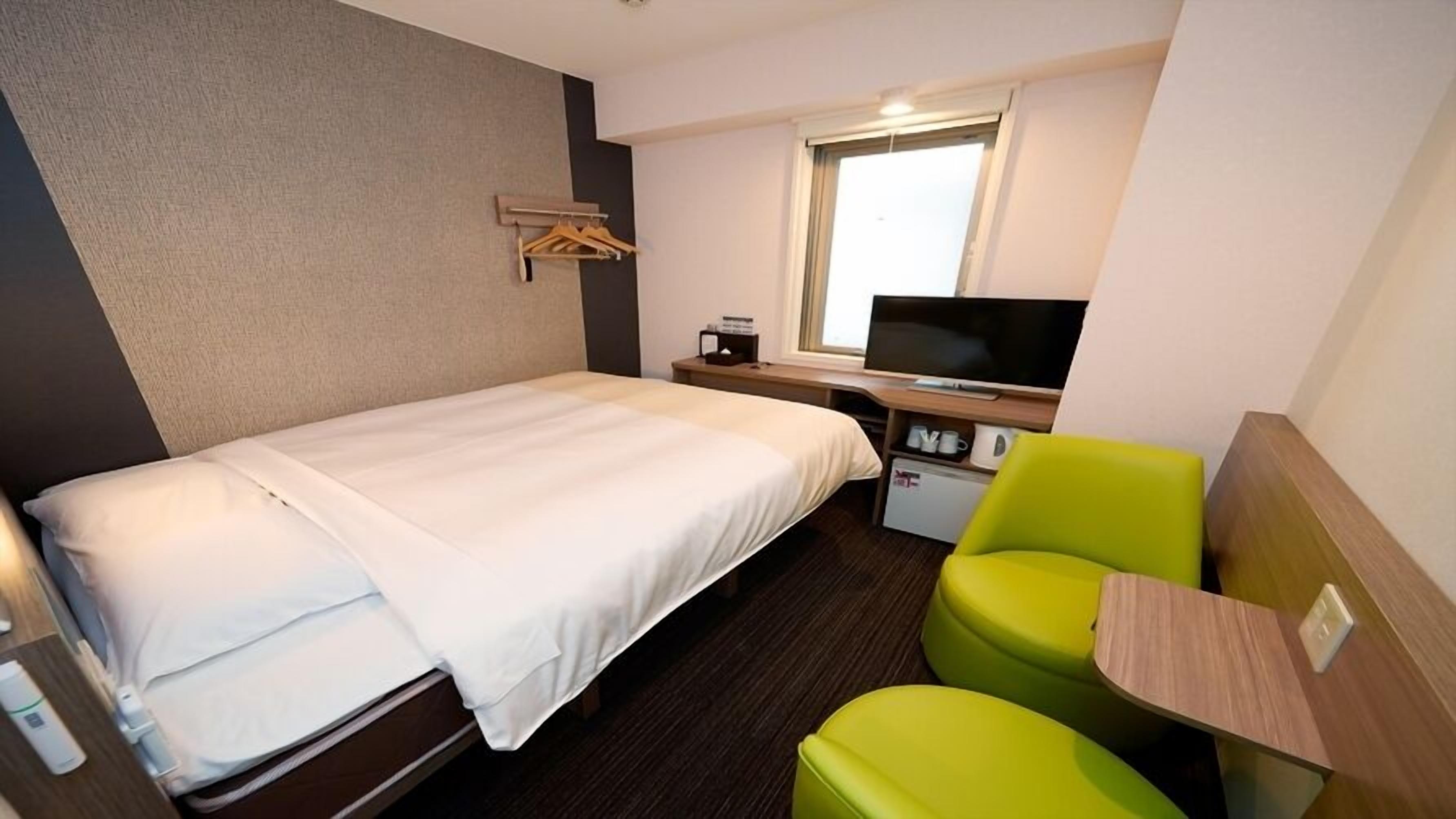 Ein Hotelzimmer mit einem Bett, einem Schreibtisch mit Fernseher, einer kleinen Küchenzeile und einem leuchtend grünen Stuhl.
