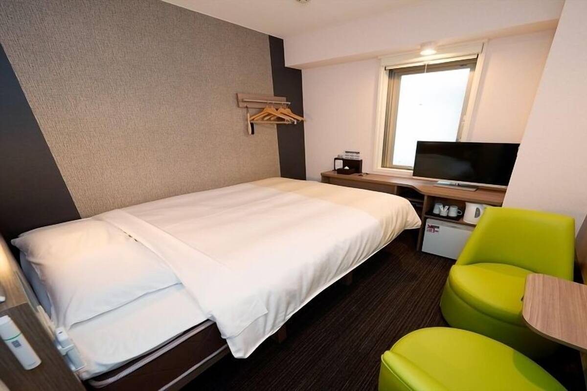 Une chambre d’hôtel avec un lit, un bureau avec une télévision, une petite kitchenette et une chaise vert vif.