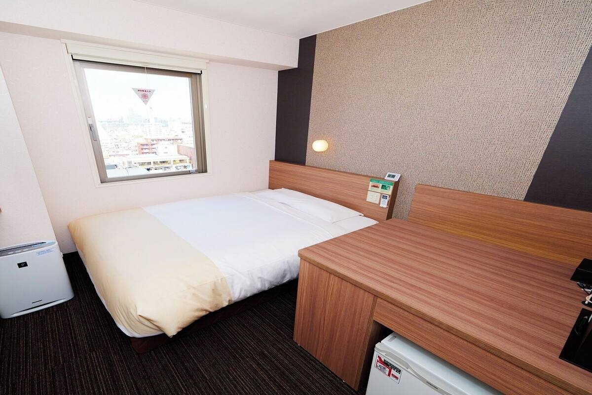 Une chambre d’hôtel équipée d’un lit, d’un bureau, d’une télévision, d’un radiateur et d’une fenêtre donnant sur la ville.